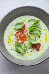 Bowl of chilled cucumber soup topped with fresh cucumber slices, red cherry tomatoes and green basil, middle closeup, vertical shot