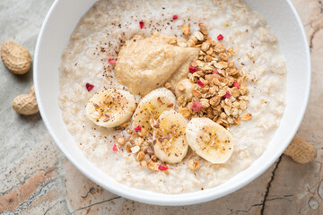 Oatmeal served with peanut butter, fresh banana and granola in a white bowl, horizontal shot, middle close-up, elevated view