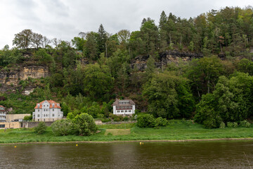 Obraz premium A house is on a hill with a river in the background. The house is white and has a red roof. The hill is covered in trees and the river is calm
