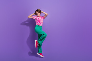 Full length photo of gorgeous pretty girl wear stylish t-shirt green pants flying hold hands on head isolated on purple color background