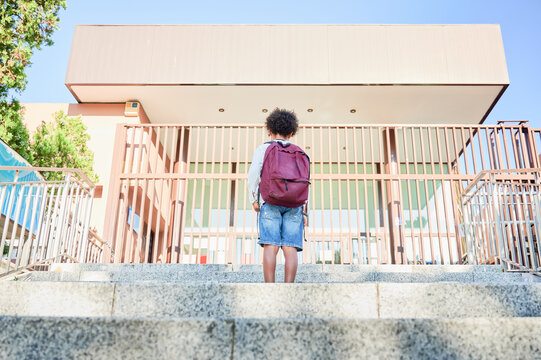 Back to school, beginning of the new primary school year. A boy with curly hair wearing shorts walks resignedly towards the school entrance, carrying his backpack.