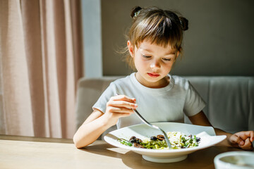 Little girl sitting at a table, savoring vegetarian pasta with green pesto and black olives