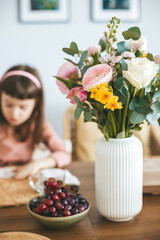 A floral bouquet in a white vase, with a little girl in the background and a bowl of mixed fruits including grapes and cherries