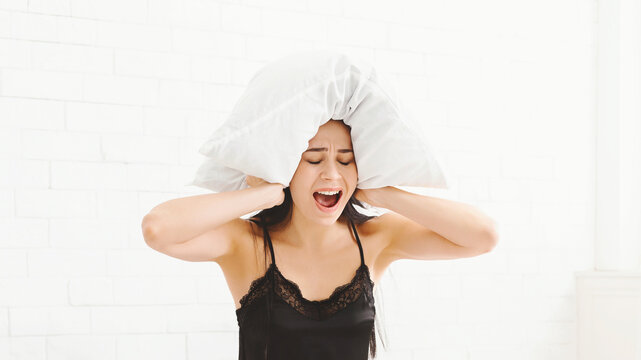 A young woman is sitting on her bed with a distressed expression, screaming while holding a pillow over her head. She looks frustrated or overwhelmed. - Powered by Adobe