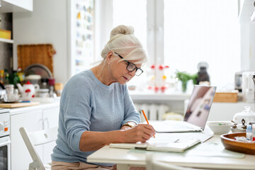 Senior woman with laptop and notebook sitting at the table in the kitchen
