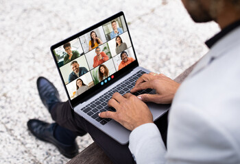 Black man sits on a bench outdoors, using his laptop to participate in a video conference meeting....
