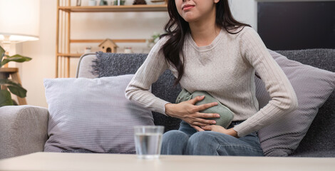 Young Woman Experiencing Menstrual Cramps at Home, Holding Heating Pad for Pain Relief, Sitting on Couch in Comfortable Living Room