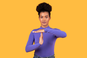 A young woman with dark curly hair is standing against a yellow background. She is wearing a purple long-sleeved shirt and is making the time out hand gesture with her hands.