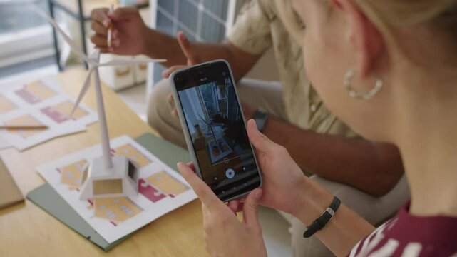 Handheld shot of woman filming video on smartphone how man explaining work of wind turbine using plastic model for their blog about alternative energy