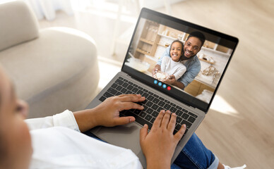 Black woman mother sits on a couch and uses a laptop to video chat with their family. The person on the laptop screen is a man and a young girl, both smiling and looking happy.