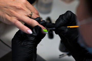 A professional manicurist in black gloves meticulously applies intricate green nail art to a...
