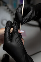 A manicurist in black gloves meticulously detailing a client’s nails with a wooden stick, highlighting precision and expert care in a salon environment.