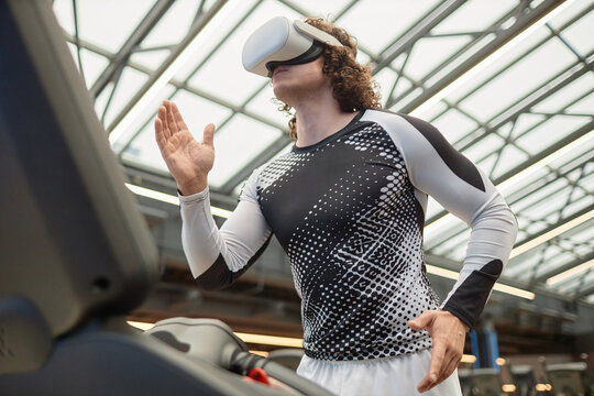 Action side view of muscular young man running on treadmill with VR headset in futuristic gym copy space
