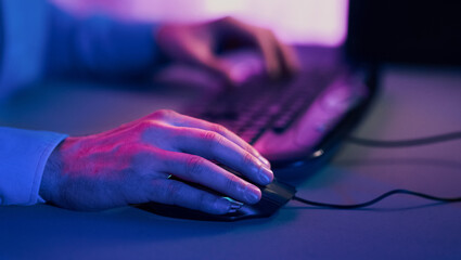 A persons hand is shown resting on a computer mouse, which is connected to a computer. The room is lit with blue and purple hues, creating a dramatic and colorful effect