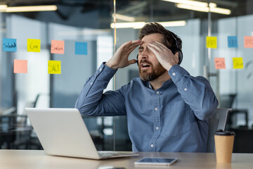 Shocked young man working in the office at the laptop, sitting at the desk and holding his head...