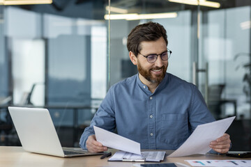 Smiling young man businessman working in office with documents and laptop, holding and reviewing papers
