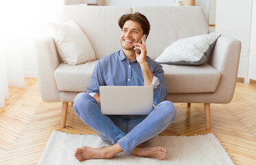 A man sits on the floor in front of a couch, using a laptop and talking on his phone.