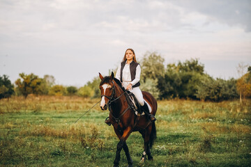 Young beautiful blonde woman jockey rides a brown horse in a meadow at sunset in summer. Preparing...