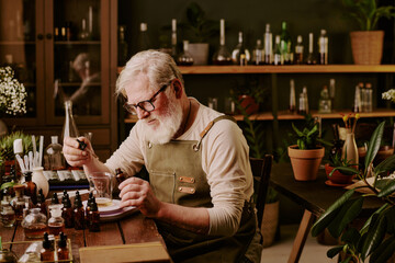 Mature perfumer dressed in suede apron with leather elements adding extra essential oil into aroma liquid in glass flask