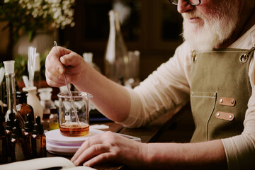Senior perfumer sitting at table with brown glass bottles of essential oils and mixing cognac shade essences with help of long glass stick