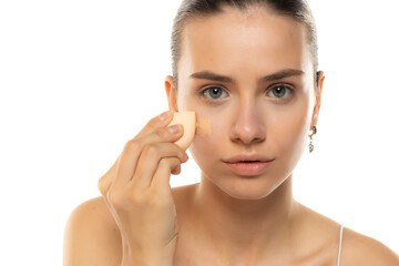 Portrait of a beautiful young woman applying makeup foundation with a sponge on a white studio background