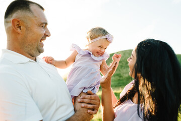 Daughter hugging parents on nature. Mom, dad and girl toddler, walk in the grass. Happy young family spending time together, outside, on vacation, outdoors. The concept of family holiday.
