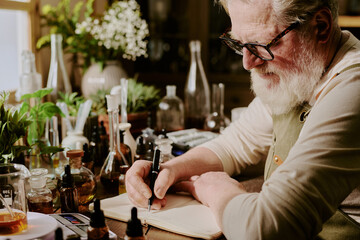 Grey-haired mature man sitting at his perfume work place and making personal notes about specific...