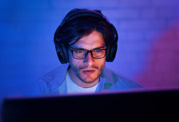 A young man wearing glasses and headphones sits in front of a computer screen in a dimly lit room....