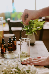 Female hands strewing dried crushed petals of daisy into transparent and clean liquid in long glass flask
