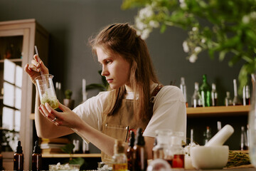 Young female perfumer sitting at work desk with perfume craft items and admiring at golden liquid...