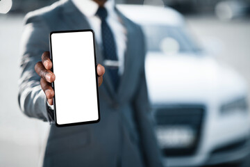 A man in a suit stands in front of a white car, holding out a smartphone with a blank white screen towards the camera, cropped