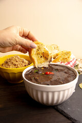 Hand with bread slice inside a black bean creamy soup with fresh herbs and pepper