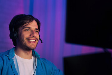 A young man smiles while looking off to the side. He is wearing a blue shirt and a headset. He is sitting in front of a computer with a purple and blue backdrop.