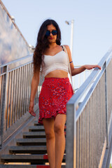 Stylish young woman in sunglasses descends a metal staircase in a vibrant red floral skirt and a...
