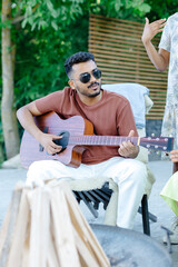 Multiracial group of people, indian man playing guitar and friends eating dinner and drinking wine during party in the picnic