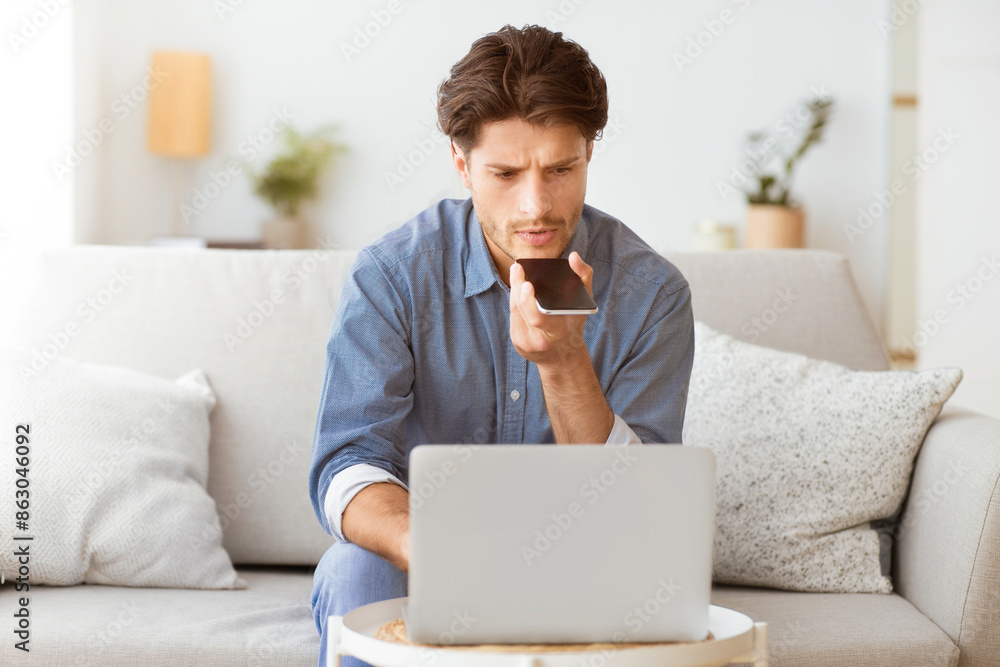 Poster A man sits on a couch in his home and uses the voice assistant on his smartphone while working on a laptop.