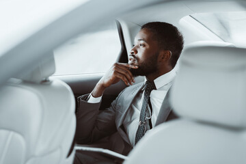 A black man in a grey suit with a white shirt and a dark tie sits in the backseat of a car looking...