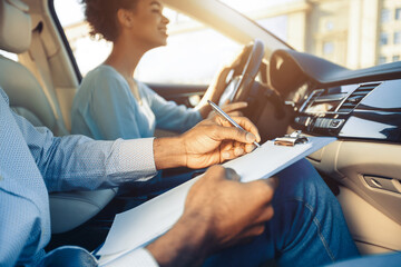 A driving instructor sits in the passenger seat of a car, holding a clipboard and pen, evaluating a student who is behind the wheel. Instructor appears to be taking notes on the students performance