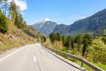 Fototapeta premium Empty winding mountain road through a forested valley in Switzerland on a clear spring day. Agricultural buildings along the road are visible in distance.