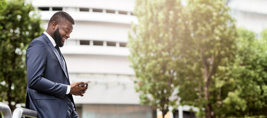 Attractive happy businessman is texting on cell phone near modern office building, side view with copy space. Business, technology and communication concept