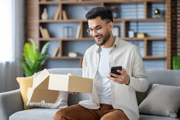 Muslim smiling young man in casual clothes and glasses is sitting on the sofa at home, holding a mobile phone and looking into an open box of a package he received by mail