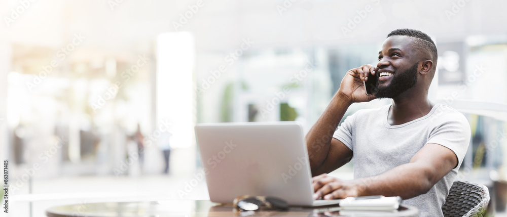 Poster A man sits at a table with a laptop open, smiling and talking on his phone.