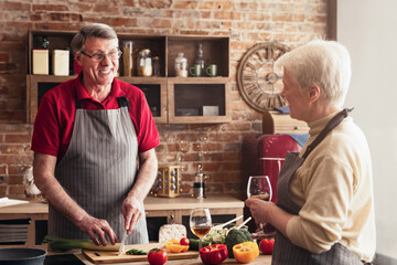 A senior couple is preparing food together in a home kitchen. The man is wearing a red shirt and a grey apron, while the woman wears a cream sweater and a black and white striped apron
