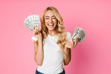 A young woman with blonde hair and a white t-shirt is smiling excitedly while holding US dollar bills in both hands. She is standing against a pink background, looking happy and successful.