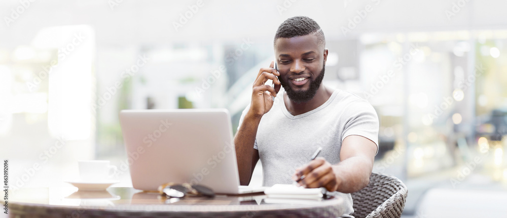 Poster a man with a beard sits at a table outdoors, talking on a phone while working on a laptop and taking