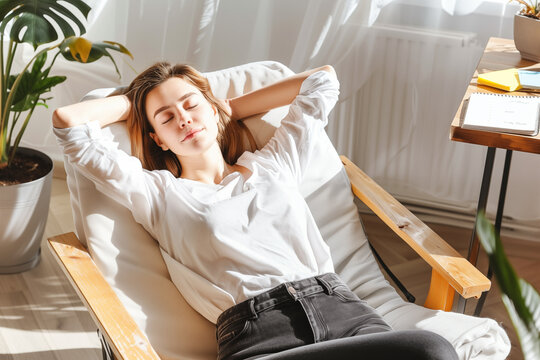 Young female relaxing on chair with arms behind head, sunlight through window