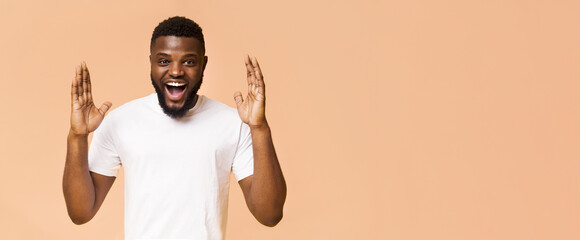 A young black man wearing a white t-shirt smiles and looks shocked while raising his hands in the...