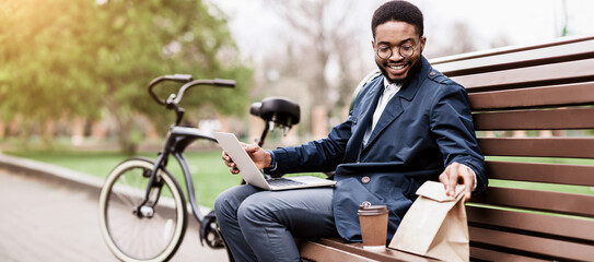 Black man sits on a park bench using his laptop, his bicycle nearby. The setting is calm and professional, reflecting a modern approach to work in a natural environment.