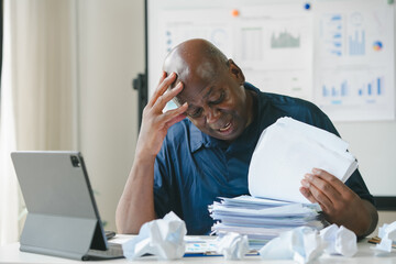 Man frustrated with paperwork and reports at desk, surrounded by crumpled paper. Laptop on, charts...