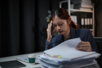 Stressed businesswoman overwhelmed with work documents, holding her head while surrounded by...